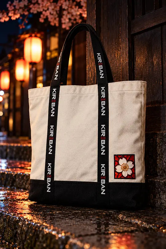 A canvas tote with black KIRIBAN straps and a small blossom patch, on stone steps at night.